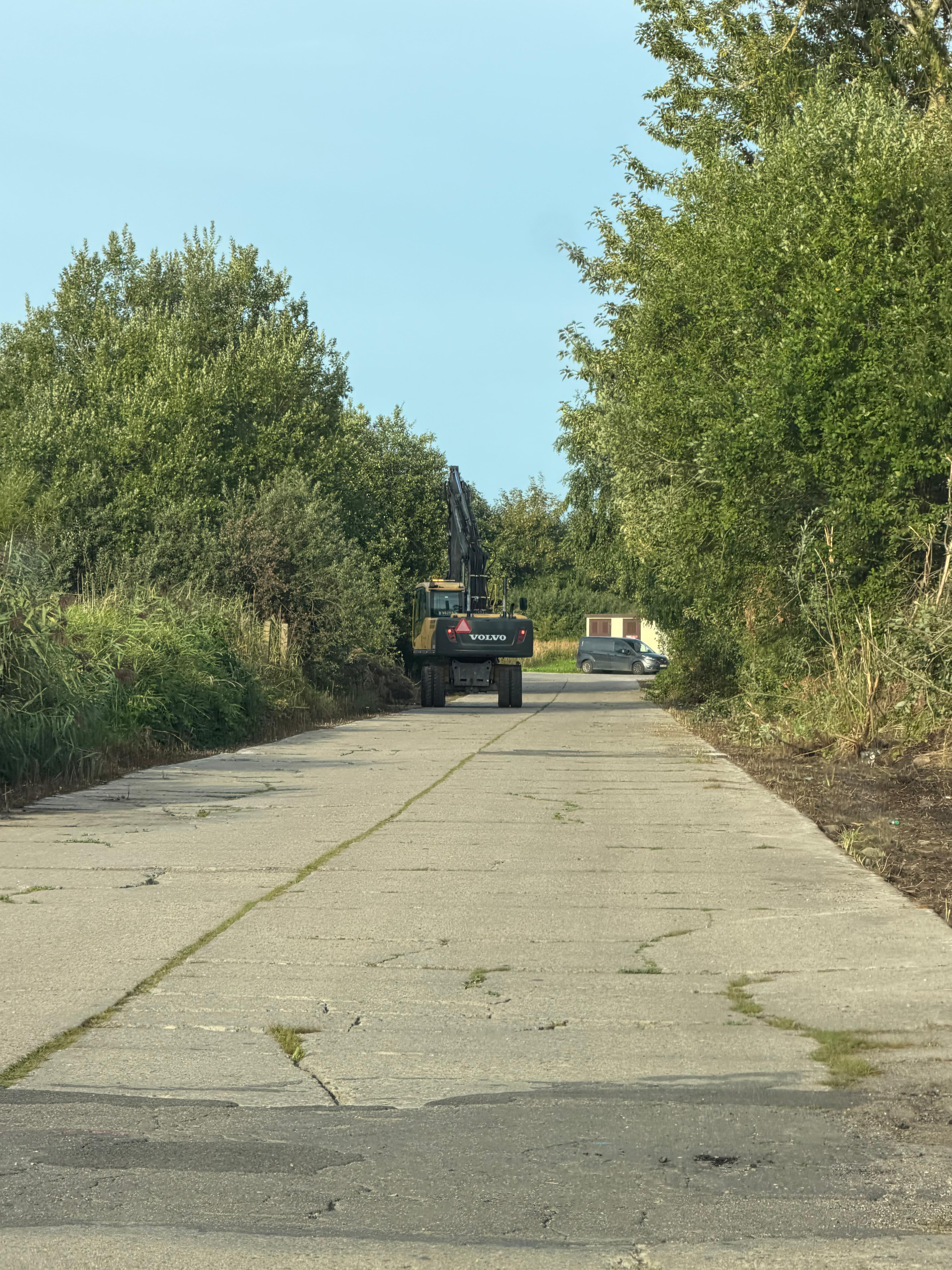 Koparka Volvo na betonowej drodze otoczonej drzewami i krzewami w słoneczny dzień Droga betonowa zarośnięta trawą, prowadząca w głąb zieleni. Na końcu drogi koparka Volvo i biały van. Niebo błękitne, słoneczny dzień.