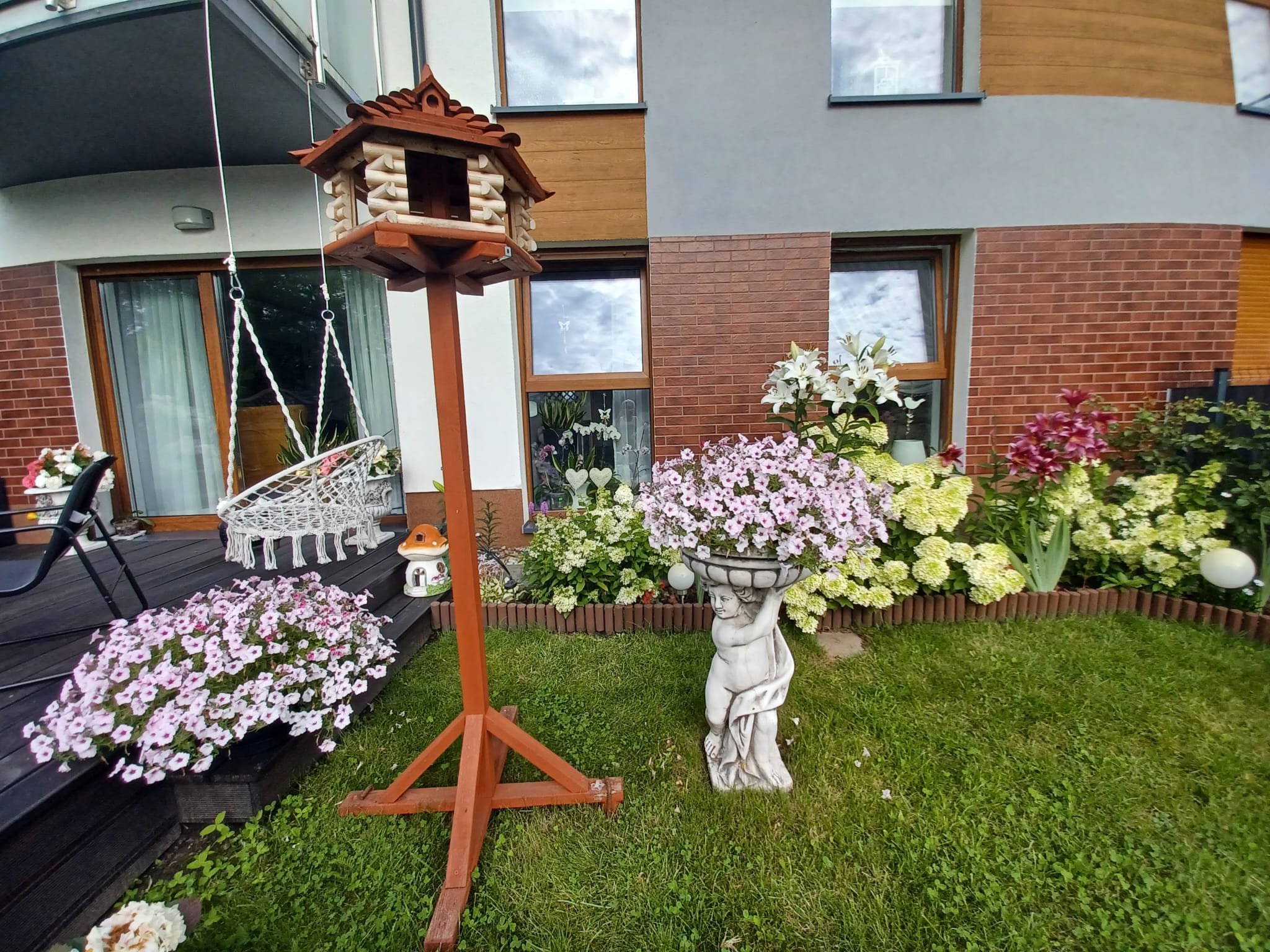 Karmnik dla ptaków i kwiaty w ogrodzie przed domem Drewniany karmnik dla ptaków na tle nowoczesnego budynku mieszkalnego z balkonem i ogrodem. W ogrodzie kwitną hortensje, lilie i petunie, a na trawniku stoi rzeźba anioła z donicą. Widoczna huśtawka makramowa.